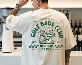 May include: Cream-colored t-shirt with a green graphic on the back that reads "Golf Dads Club." The graphic features a golf ball character with sunglasses and golf clubs. The text "Best Dad by Par" is also included.