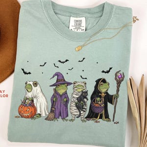 May include: A bay-colored short-sleeve t-shirt featuring a graphic of four frogs dressed in Halloween costumes: one as a ghost with a pumpkin bucket, one as a witch with a broom, one as a mummy holding a black cat, and one as a wizard with a staff.