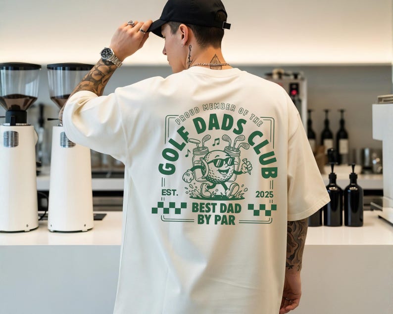 May include: Cream-colored t-shirt with a green graphic on the back that reads "Golf Dads Club." The graphic features a golf ball character with sunglasses and golf clubs. The text "Best Dad by Par" is also included.