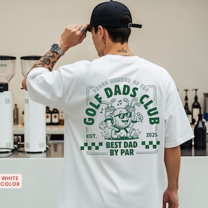 May include: White t-shirt with a green graphic that reads "GOLF DADS CLUB" and "BEST DAD BY PAR". The graphic features a golf ball character wearing sunglasses and holding golf clubs. The shirt also has the text "EST. 2025".