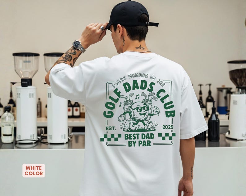 May include: White t-shirt with a green graphic that reads "GOLF DADS CLUB" and "BEST DAD BY PAR". The graphic features a golf ball character wearing sunglasses and holding golf clubs. The shirt also has the text "EST. 2025".