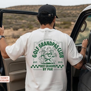 May include: Ivory-colored t-shirt with a green graphic that reads "GOLF GRANDPAS CLUB" and "BEST GRANDPA BY PAR." The design features a golf ball character with sunglasses and golf clubs. The shirt also has the text "EST. 2025."