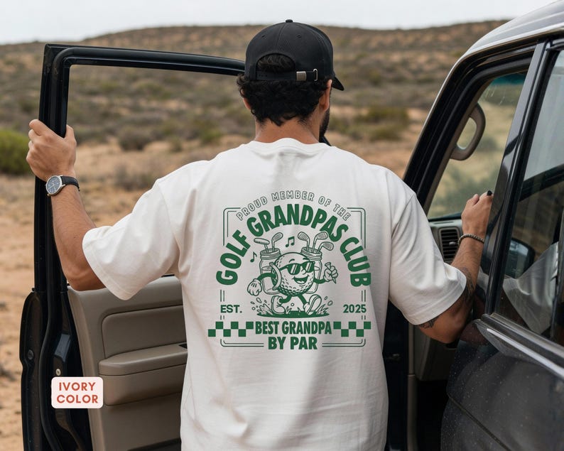 May include: Ivory-colored t-shirt with a green graphic that reads "GOLF GRANDPAS CLUB" and "BEST GRANDPA BY PAR." The design features a golf ball character with sunglasses and golf clubs. The shirt also has the text "EST. 2025."
