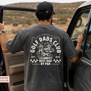 May include: A dark gray t-shirt with a graphic on the back that reads "Proud Member of the Golf Dads Club". The graphic features a cartoon golf ball character with sunglasses, golf clubs, and the text "Best Dad by Par".