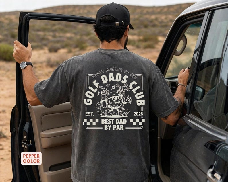 May include: A dark gray t-shirt with a graphic on the back that reads "Proud Member of the Golf Dads Club". The graphic features a cartoon golf ball character with sunglasses, golf clubs, and the text "Best Dad by Par".