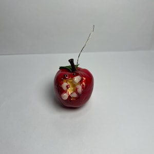 May include: A glossy red apple ornament featuring a bite mark that reveals a pale yellow interior and several white teeth. The apple has a dark brown stem, a small green leaf, and a thin silver hanging loop attached to the top of the fruit.