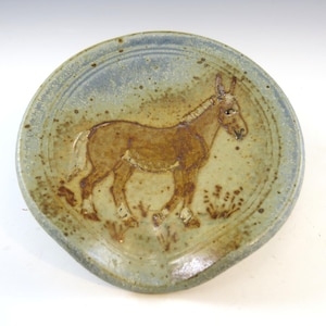 May include: A ceramic spoon rest with a hand-painted donkey design. The plate is round with a light blue and tan speckled glaze. The donkey is brown with a tan belly and is standing on a patch of grass.