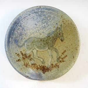 May include: A circular ceramic plate featuring a raised relief of a donkey. The plate has a speckled, muted colour palette of blue, green, and brown. The donkey is shown in motion, with details of grass and text near its feet.