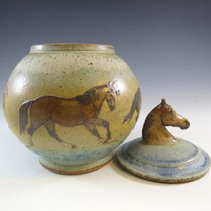 May include: A ceramic jar with a lid featuring a horse head handle. The jar is round with a light green and brown glaze, and a brown horse illustration. The lid is light blue and brown.