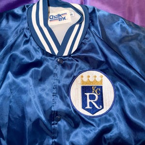 May include: A royal blue satin jacket with a white and navy striped collar and cuffs. The jacket features a circular patch with a gold crown, the letters "KC", and the letter "R". The tag reads "Chalk Line".