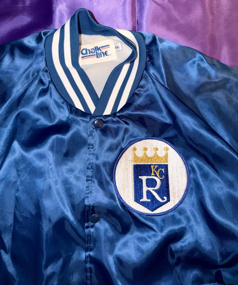 May include: A royal blue satin jacket with a white and navy striped collar and cuffs. The jacket features a circular patch with a gold crown, the letters "KC", and the letter "R". The tag reads "Chalk Line".