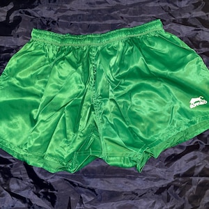 May include: Bright green athletic shorts with a shiny, satin-like finish. The shorts have an elastic waistband and a white logo of a buffalo on the lower right leg. The shorts are likely vintage or retro style.