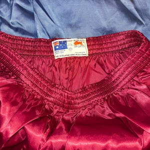 Puede incluir: Primer plano de unos pantalones cortos deportivos granates con una textura brillante, similar al sat&eacute;n. La cintura es el&aacute;stica y fruncida. Se ve una etiqueta con texto y una bandera. Los pantalones cortos son 100% nailon.