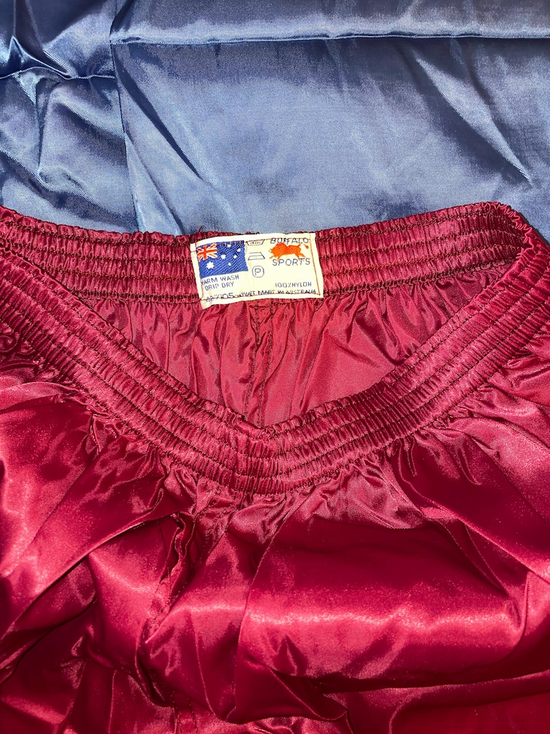 Puede incluir: Primer plano de unos pantalones cortos deportivos granates con una textura brillante, similar al sat&eacute;n. La cintura es el&aacute;stica y fruncida. Se ve una etiqueta con texto y una bandera. Los pantalones cortos son 100% nailon.