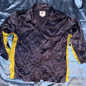 May include: A navy blue windbreaker jacket with a shiny finish, featuring bright yellow side panels and long sleeves. The jacket has a snap-button front closure, a collar, and a small yellow Nike swoosh logo on the chest. A Nike label is at the neck.