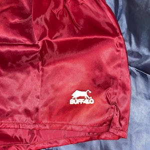 Puede incluir: Pantalones cortos de sat&eacute;n burdeos con el logo blanco "BUFFALO" con la silueta de un b&uacute;falo. Los pantalones cortos tienen cintura con cord&oacute;n y abertura en el dobladillo. Hechos de un material brillante y ligero.