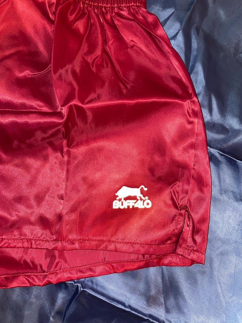 Puede incluir: Pantalones cortos de sat&eacute;n burdeos con el logo blanco "BUFFALO" con la silueta de un b&uacute;falo. Los pantalones cortos tienen cintura con cord&oacute;n y abertura en el dobladillo. Hechos de un material brillante y ligero.