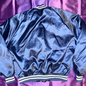 May include: A navy blue satin bomber jacket with white and navy striped cuffs and hem. The jacket has a shiny, smooth texture and a classic bomber jacket design. The collar has a white stripe.