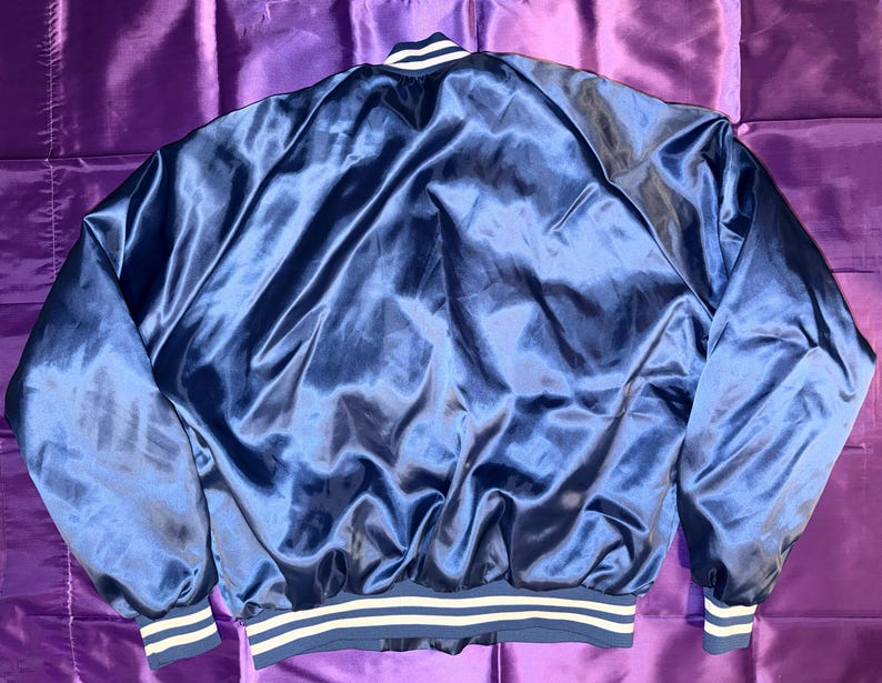 May include: A navy blue satin bomber jacket with white and navy striped cuffs and hem. The jacket has a shiny, smooth texture and a classic bomber jacket design. The collar has a white stripe.