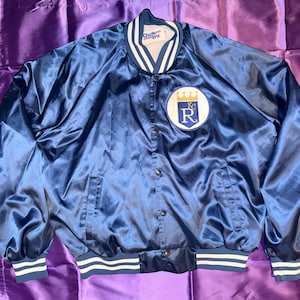 May include: A navy blue satin jacket with white and blue striped trim on the cuffs and waist. The jacket features a patch with a white background, a blue letter "R", and a gold crown. The jacket has a snap front closure.