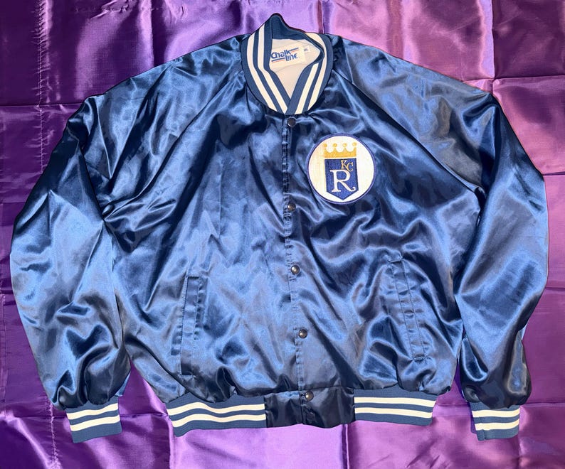 May include: A navy blue satin jacket with white and blue striped trim on the cuffs and waist. The jacket features a patch with a white background, a blue letter "R", and a gold crown. The jacket has a snap front closure.