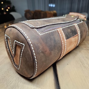 Op de afbeelding: Een bruine leren cilindrische tas met een ritssluiting en witte stiksels. De tas heeft een bruine leren patch met een witte "V" aan de voorkant.