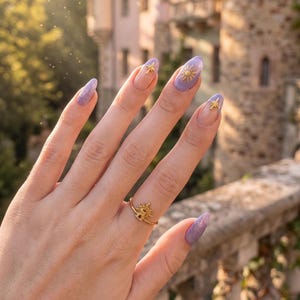 May include: A hand with long, oval nails painted in a light purple glitter polish. The nails are decorated with gold sun and star charms. A gold ring with a sun design is on the ring finger.