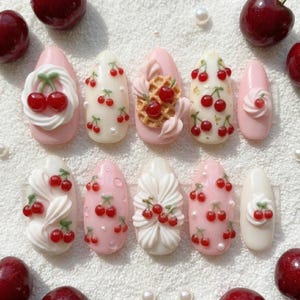 May include: A set of ten almond-shaped artificial nails with cherry designs. The nails feature various designs, including cherries, waffle patterns, and pearl accents, in shades of pink, white, and red. Fresh cherries are scattered around the nails.
