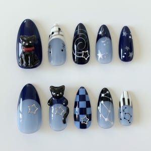 May include: A set of ten press-on nails in various designs and colors. The nails feature a dark blue base with light blue accents, cat and star embellishments, and patterns including musical notes, swirls, and checkered designs. The nails are oval-shaped.