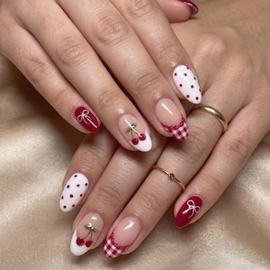 May include: A close-up of a hand with almond-shaped nails. The nails feature a variety of designs, including red polka dots, cherries, gingham, and bows. The color palette includes red, white, and nude tones. Gold rings adorn the fingers.