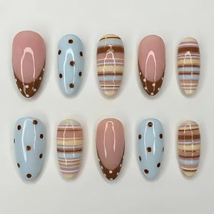 May include: A set of ten press-on nails featuring almond-shaped tips with various designs. Patterns include light blue with brown polka dots, horizontal stripes in shades of brown, cream, and blue, and solid pink with brown tips and white dots.