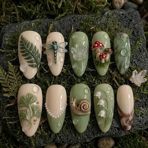 May include: A collection of ten almond-shaped, decorated artificial nails. Designs include a fern, dragonfly, mushrooms, and a snail. Colors are primarily cream, green, and brown, with clear accents. The nails are arranged on a dark, mossy surface.