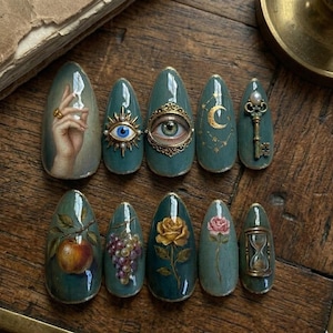 May include: A set of ten teal-colored, almond-shaped press-on nails. Each nail features a unique design, including a hand with a ring, an eye, a key, a moon, a rose, an apple, grapes, and an hourglass. The designs are painted in a vintage style.