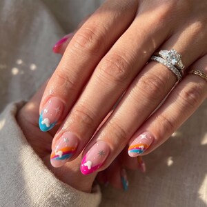 Puede incluir: Una mano con manicura con base transparente y dise&ntilde;os coloridos. Cada u&ntilde;a tiene un dise&ntilde;o diferente, incluyendo un cielo azul con una nube, un arco&iacute;ris y una estrella. Las u&ntilde;as son ovaladas y tienen un acabado brillante. La mano tambi&eacute;n tiene anillos.