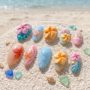 Peut inclure: Un ensemble de dix faux ongles décorés sur le thème de la plage, avec des fleurs de frangipanier en relief, des étoiles de mer, des dauphins et des motifs de vagues dans des tons bleu, rose, pêche et crème, posés sur du sable blanc.