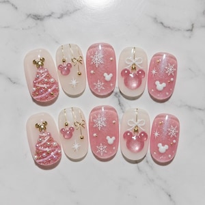 May include: A set of ten press-on nails featuring a pink and white holiday theme. Designs include a pink glitter Christmas tree with a gold Mickey head topper, pink Mickey ornaments with gold bows, white snowflakes, and small white Mickey silhouettes.