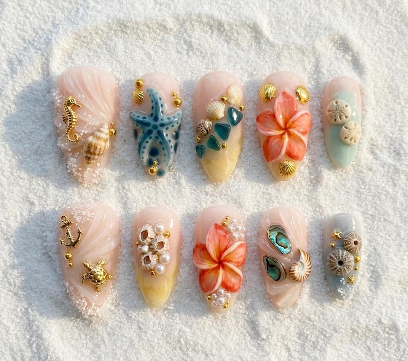 May include: A set of ten artificial nails with a beach theme. Each nail features a different design, including seahorses, starfish, shells, and flowers, with gold accents. The nails are painted in shades of pink, blue, and yellow.