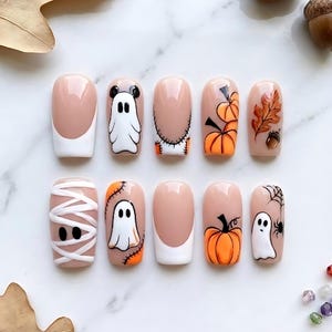 May include: A set of ten press-on nails featuring a nude base with various Halloween-themed designs including white ghosts, orange pumpkins, a mummy wrap pattern, a spider with a web, autumn leaves, acorns, and classic white French tips.