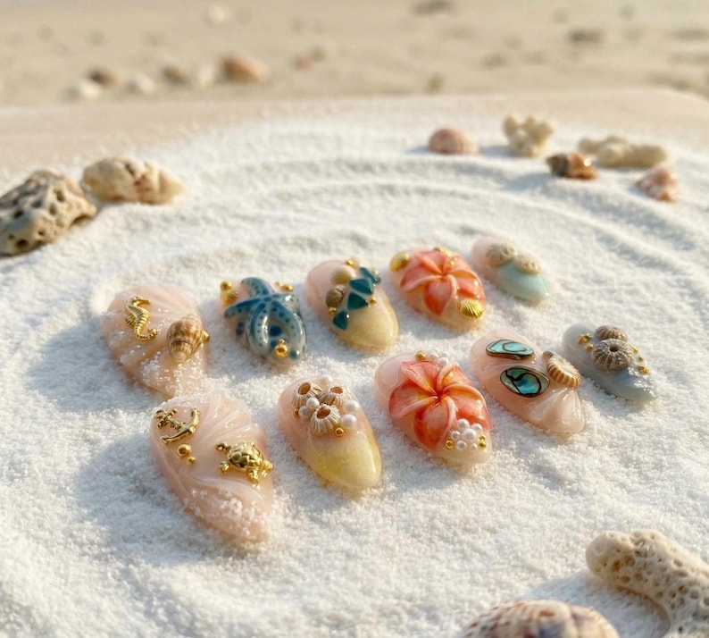 May include: A collection of ten artificial nails, each adorned with unique, beach-themed embellishments. Designs include seahorses, starfish, and floral accents, set against a backdrop of white sand. The nails feature a variety of colors, including pink, blue, and gold.