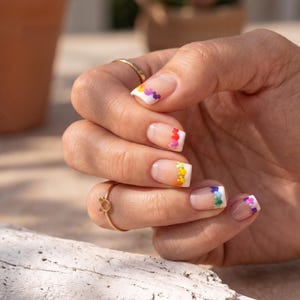 May include: Close-up of a hand with manicured nails. The nails feature a clear base with white tips, each adorned with a colorful, rainbow-themed floral design. A gold ring with a small charm is also visible.