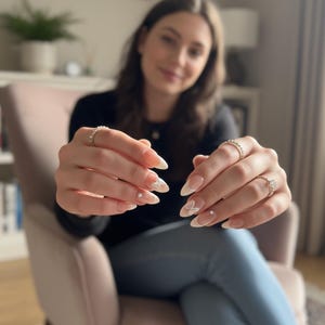 Puede incluir: Primer plano de u&ntilde;as manicuradas con un dise&ntilde;o de punta francesa blanca y peque&ntilde;os acentos de flores blancas. Las u&ntilde;as son largas y en forma de almendra. La imagen tambi&eacute;n muestra tres anillos con piedras transparentes en los dedos.