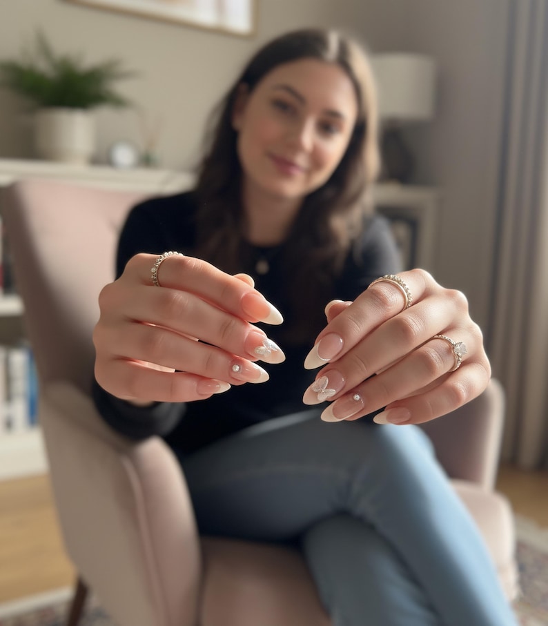 Puede incluir: Primer plano de u&ntilde;as manicuradas con un dise&ntilde;o de punta francesa blanca y peque&ntilde;os acentos de flores blancas. Las u&ntilde;as son largas y en forma de almendra. La imagen tambi&eacute;n muestra tres anillos con piedras transparentes en los dedos.