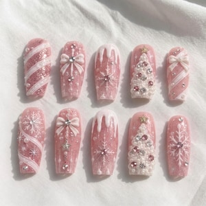 Puede incluir: Un juego de diez uñas postizas con base de purpurina rosa y detalles blancos en 3D. Los diseños incluyen rayas diagonales, lazos, glaseado que gotea, copos de nieve y árboles de Navidad decorados con pequeñas perlas blancas y brillantes.