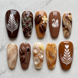 May include: A set of ten press-on nails featuring coffee-themed designs in shades of brown, cream, and tan. The nails display intricate details including 3D coffee beans, a white swan, a silver spoon, ice cubes, a lemon slice, and latte art.