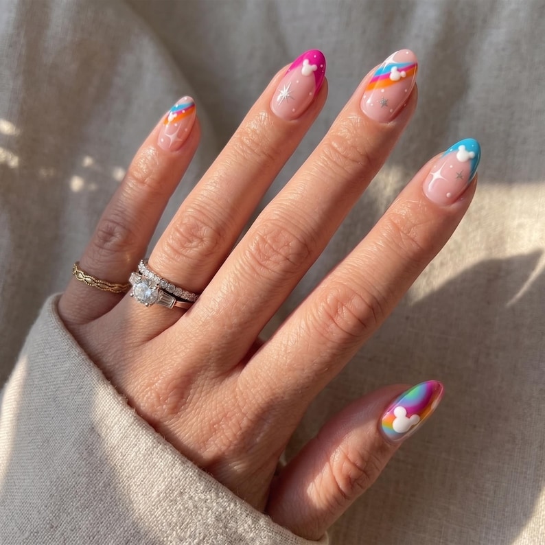 Puede incluir: Una mano con u&ntilde;as en forma de almendra con dise&ntilde;os coloridos. Las u&ntilde;as presentan rayas arco&iacute;ris, motivos de Mickey Mouse y detalles de estrellas. La mano tambi&eacute;n muestra dos anillos, uno con un diamante y otro con una banda dorada.