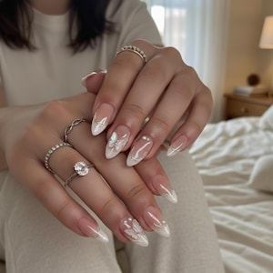 Puede incluir: Elegantes u&ntilde;as almendradas con dise&ntilde;o de manicura francesa. Las u&ntilde;as presentan una base transparente con puntas blancas, adornadas con delicados acentos de mariposas y perlas. Varios anillos plateados son visibles en los dedos.