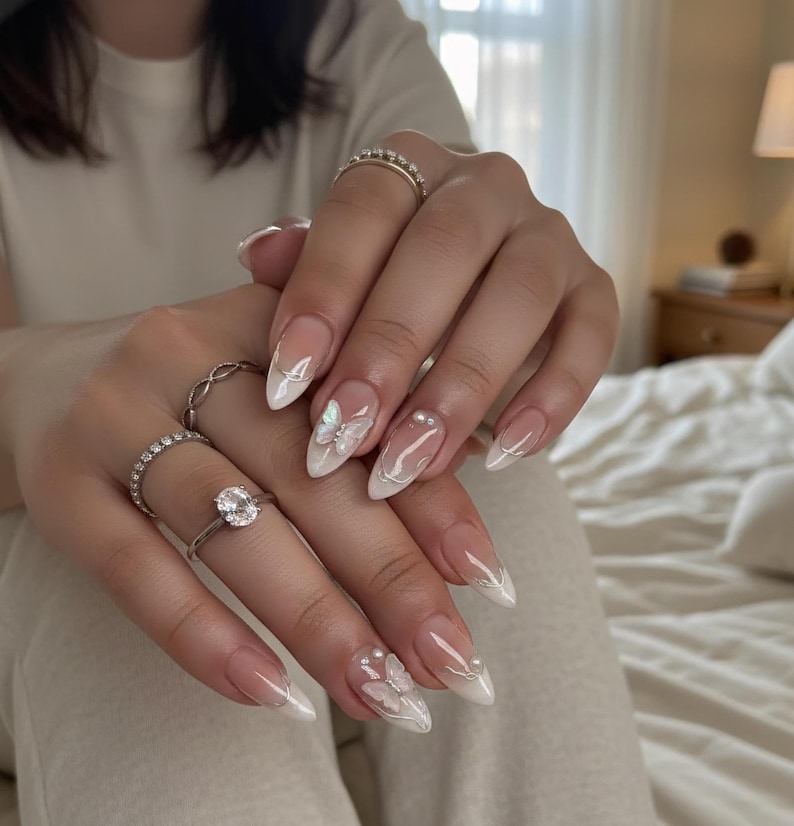 Puede incluir: Elegantes u&ntilde;as almendradas con dise&ntilde;o de manicura francesa. Las u&ntilde;as presentan una base transparente con puntas blancas, adornadas con delicados acentos de mariposas y perlas. Varios anillos plateados son visibles en los dedos.