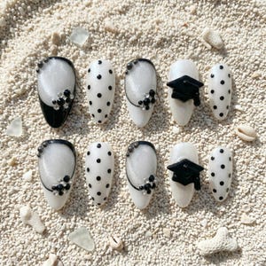 May include: A set of ten artificial nails with a white base, black polka dots, and black and silver accents. Some nails feature a black graduation cap design. The nails are arranged on a sandy surface.