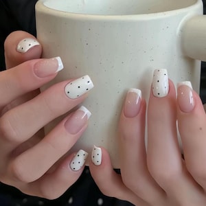 May include: A close-up of manicured nails. The nails feature a natural base with white tips and black polka dots on some nails. The nails are square-shaped and are held near a white mug.
