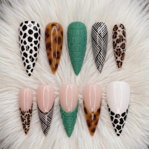 May include: A collection of ten long, pointed artificial nails. The designs include black and white spots, animal print, green crocodile texture, and black and white diagonal stripes. Some nails have a nude base with detailed designs.